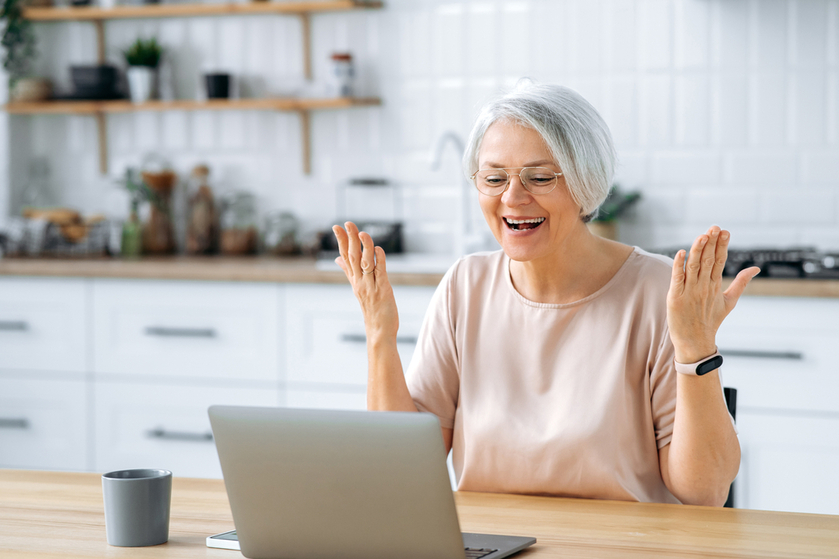 Cheerful excited middle aged modern caucasian gray haired woman, with glasses, sits at home kitchen at a table with laptop, works remotely on a project, manages finances, rejoices at result, smiles