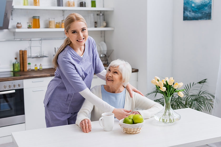 Young nurse smiling at camera while embracing happy aged woman kitchen